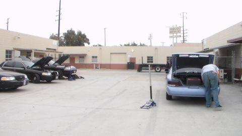 2003 Ford Crown Victoria - light blue sedan sits with its trunk open as a person leans inside, parked in a concrete lot near a metals-yard sign and other cars.