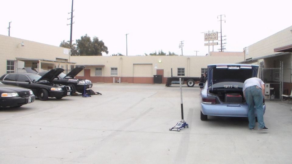 Light blue sedan with open trunk and a person leaning in, parked in an industrial lot