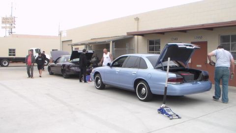 2003 Ford Crown Victoria - Several people stand around a dark sedan and a light blue sedan, both with trunks open, parked in front of numbered loading-dock units.