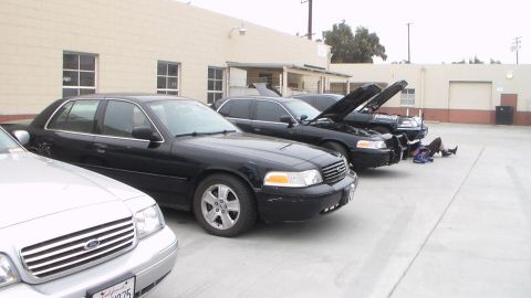 2003 Ford Crown Victoria - row of black full-size sedans parked nose-out beside a tan building, one with its hood raised and a person working underneath.