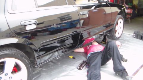 2003 Ford Crown Victoria - person in a red shirt lies on a plastic sheet working beneath a glossy black sedan with red brake calipers and a Ford oval, inside a shop.