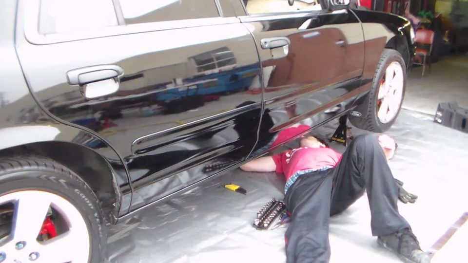 Person in red shirt lying under a black sedan with a Ford oval on the door area, in a shop