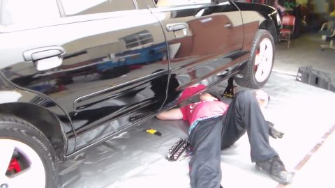 2003 Ford Crown Victoria - person in a red shirt works underneath the rocker of a glossy black sedan bearing a Ford oval badge, on a plastic sheet in a shop.