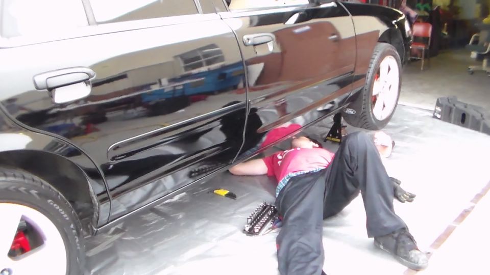 Person in red shirt working under the side of a glossy black sedan with a Ford oval badge