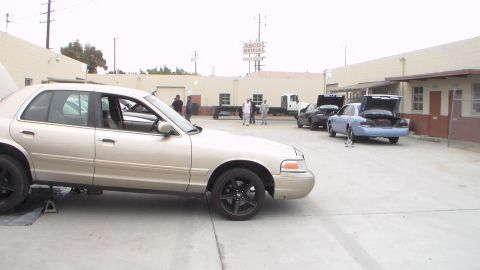 2003 Ford Crown Victoria - Photo of a gold Ford Crown Victoria parked in a shop lot with people and vehicles in background, not a technical document.