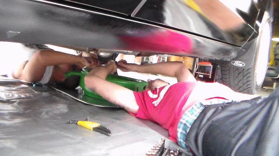 Two people lying under a black car working with a green floor jack