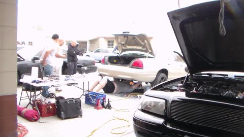 2003 Ford Crown Victoria - black car sits with its hood up beside a silver Ford Crown Victoria while people work amid tools and crates in a garage lot.
