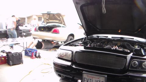 2003 Ford Crown Victoria - black sedan with a billet grille and raised hood shows a California plate reading EVTLVIC, parked beside a silver Crown Victoria.
