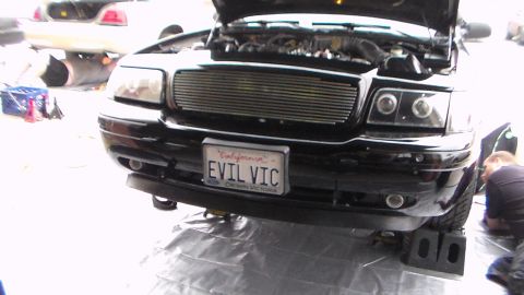 2003 Ford Crown Victoria - Photo of a black Ford Crown Victoria with 'EVIL VIC' plate and open hood in a shop, not a technical document.