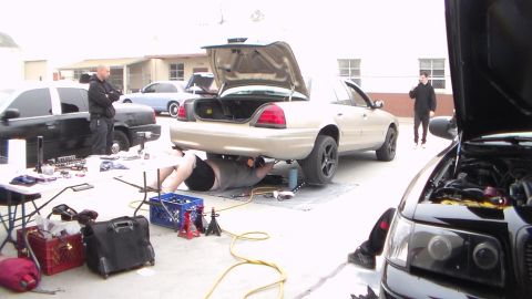 2003 Ford Crown Victoria - person lies beneath a silver Ford Crown Victoria with its trunk open while tools and crates surround the work area in a garage lot.
