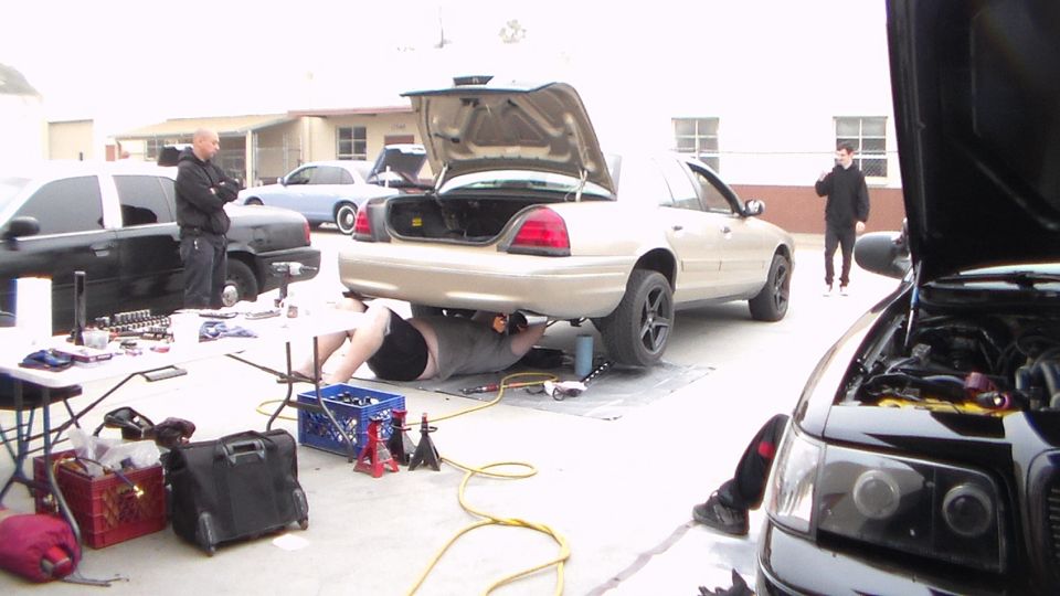 People working under a silver Crown Victoria with trunk and hoods open in a lot