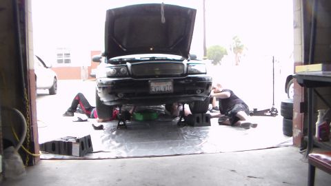 2003 Ford Crown Victoria - Two people lie working beneath a black car with its hood up in an open garage doorway over a silver tarp, tires stacked at right.