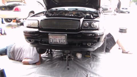 2003 Ford Crown Victoria - black car with a billet grille and open hood shows a California EVIL VIC plate as two people work on the ground beside the front.