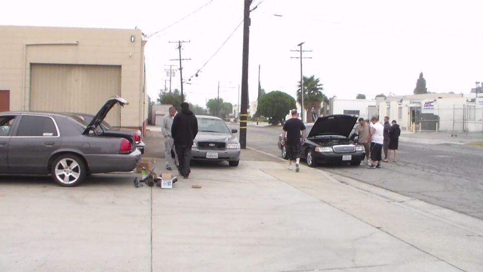 Several cars parked along a street with hoods and trunks open