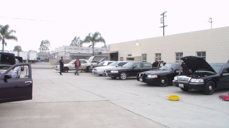 Row of parked cars with open hoods and people in a lot