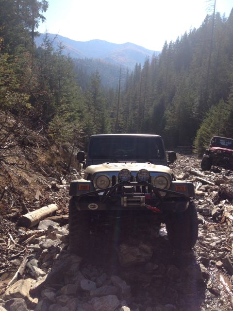 2005 Jeep Wrangler - A tan Jeep Wrangler is navigating a rocky, wooded trail. The front of the vehicle is visible, showing aftermarket lights and a winch.