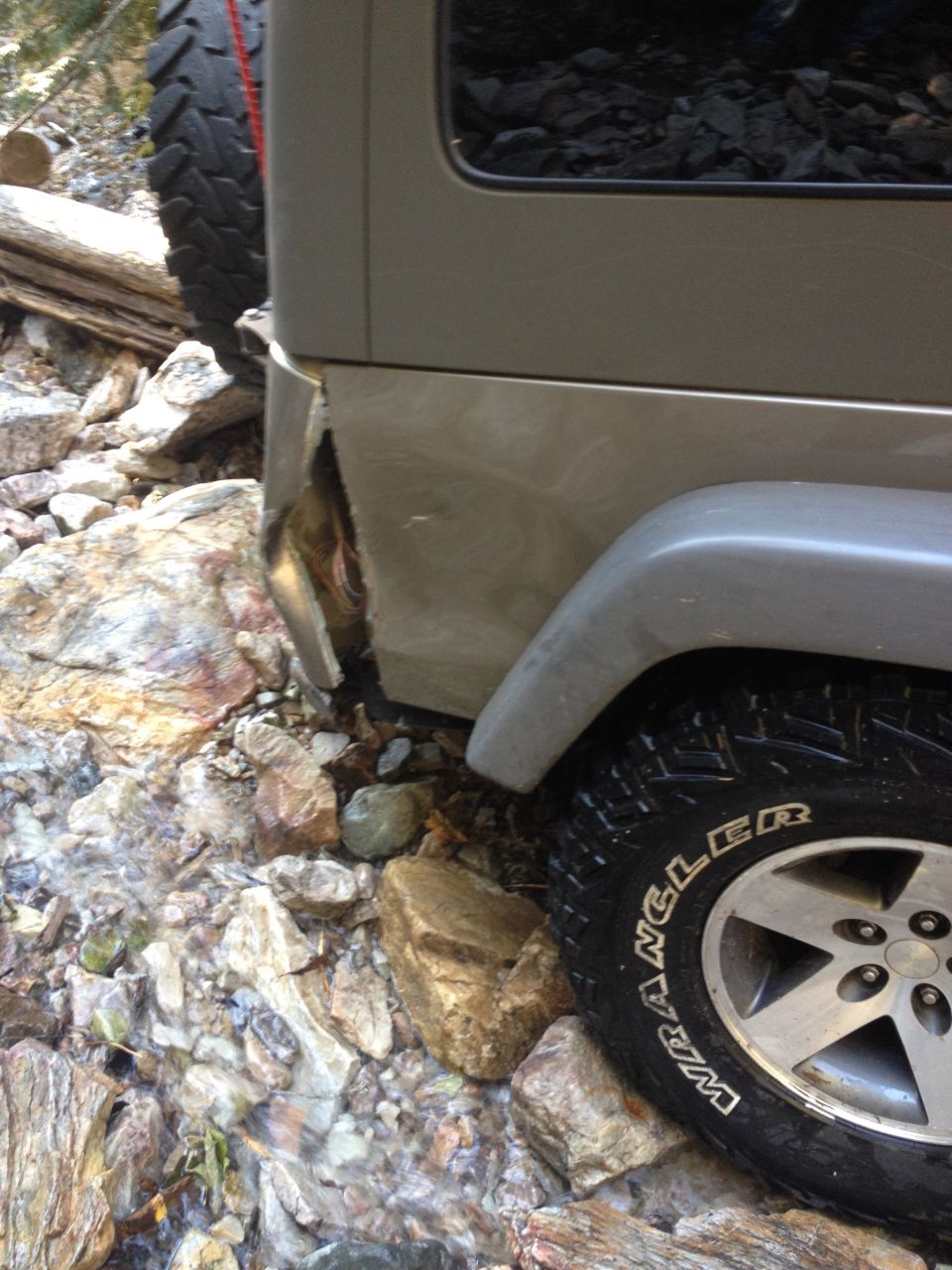 Damaged rear corner of a silver Jeep Wrangler in a rocky stream.