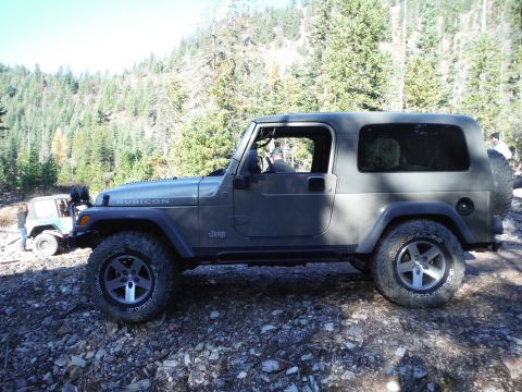 2005 Jeep Wrangler - A dark gray Jeep Wrangler Rubicon is parked on a rocky, wooded trail. Another light-colored Jeep is visible behind it.