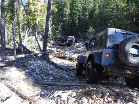 2005 Jeep Wrangler - A dark gray Jeep Wrangler is navigating a rocky, wooded trail. Another Jeep is visible further up the trail behind it.