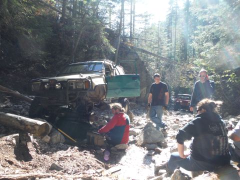 2005 Jeep Wrangler - Several people are gathered around a modified off-road vehicle stuck on a rocky, wooded trail.