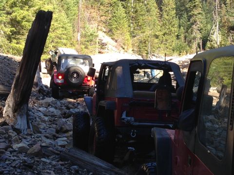 2005 Jeep Wrangler - Multiple Jeep Wranglers navigate a challenging rocky trail through a forest. The lead vehicle is red.
