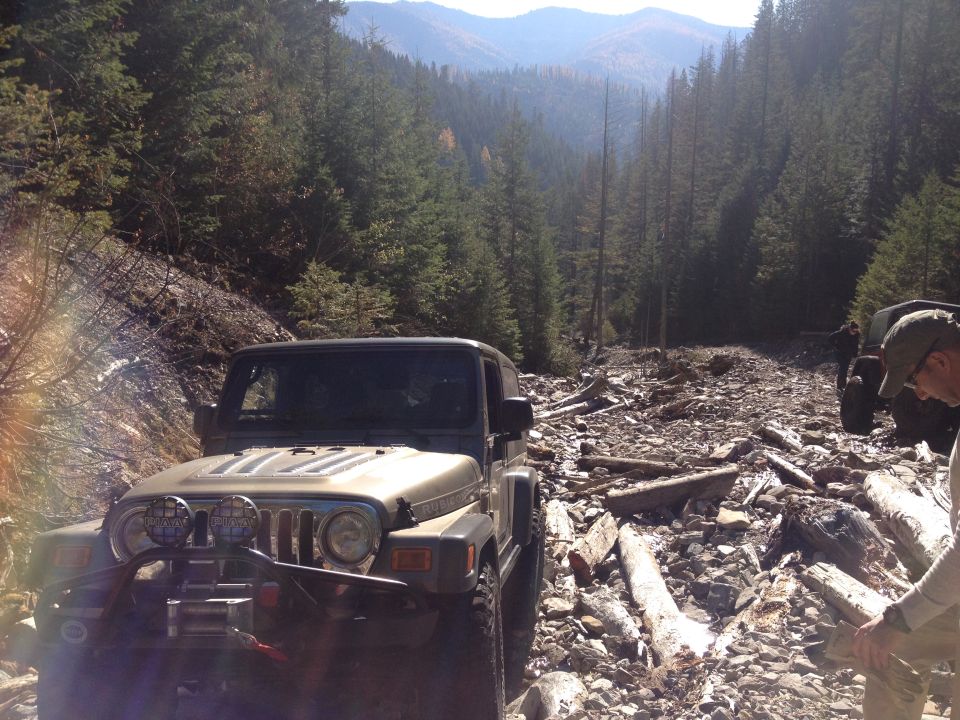 Tan Jeep Wrangler on rocky mountain trail, front bumper with winch and lights, trees and mountains in background