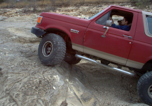 Red 1989 Ford Bronco driving over rocks, driver visible inside