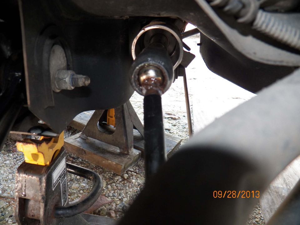 Under-car view of a ball stud end link and mount during repair, vehicle supported on jack stands over gravel