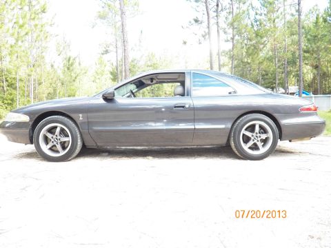 1998 Lincoln Mark VIII - Side view of a dark gray coupe with aftermarket wheels, parked on a dirt surface with trees in the background.