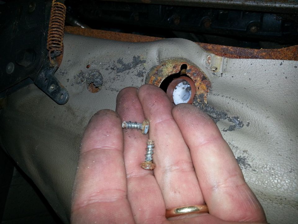 Hand holding rusty screws near a worn, tan seat bracket with a visible spring