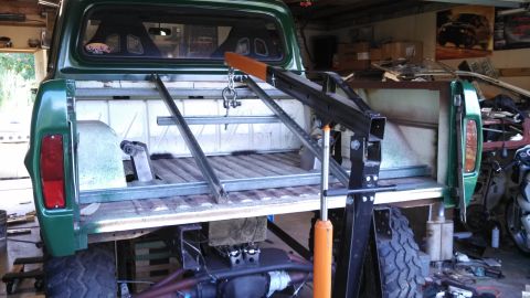 1967 Ford F-250 - A green pickup truck bed is viewed from the rear, with an engine hoist positioned over the bed and exposed engine parts below.
