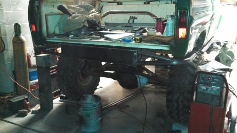 1967 Ford F-250 - Custom roll cage and large tires are fitted to the rear of a green pickup truck in a workshop.
