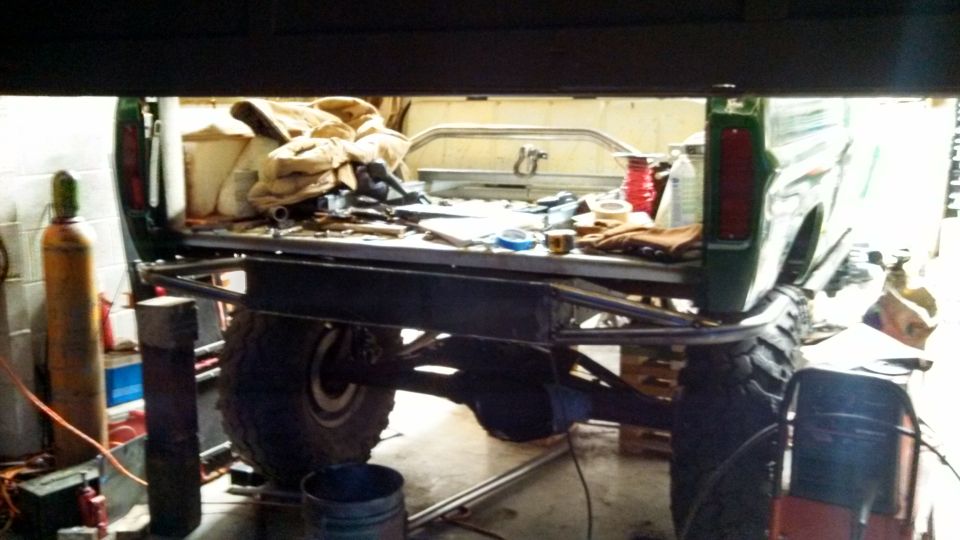 Back of a green pickup truck with a custom bumper and a cluttered bed, inside a garage