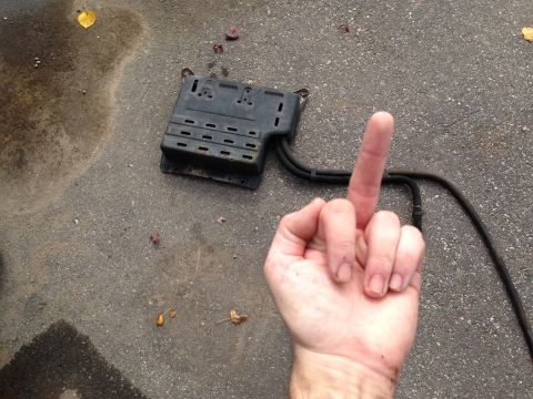 2004 Ford Crown Victoria - A black automotive part with hoses lies on asphalt next to a hand giving the middle finger, with a water stain nearby.