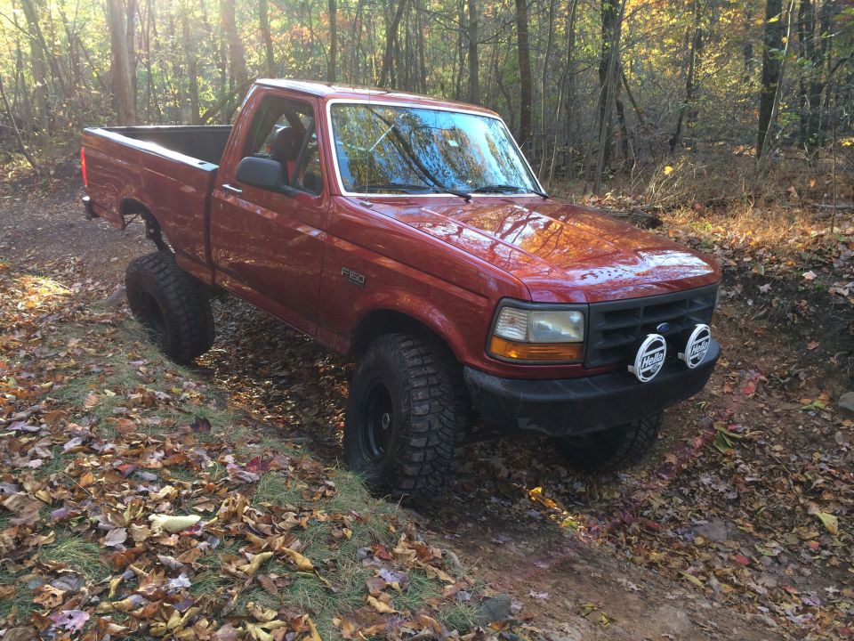 Red 1996 Ford F-150 with off-road tires on a wooded trail