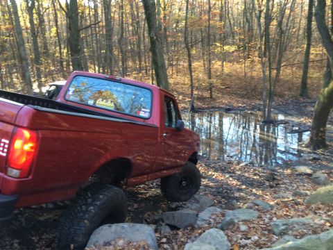 1996 Ford F-150 - A red pickup truck is shown off-roading through a wooded area with a muddy puddle and rocks.