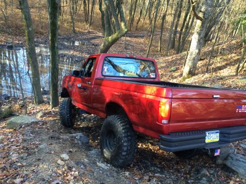 1996 Ford F-150 - A red pickup truck navigates a wooded trail with large tires and a lifted suspension.