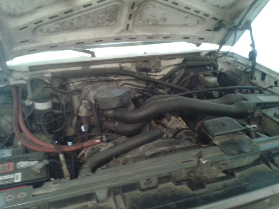 Engine bay of a white 1988 Ford Bronco showing a dusty V8 engine, hoses, air cleaner, and battery.