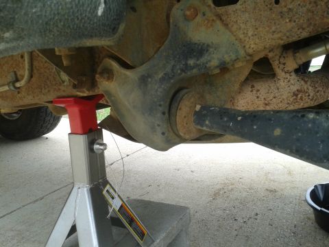 1988 Ford Bronco - The underside of a vehicle is shown, supported by a jack stand, with visible rust and a drain pan.