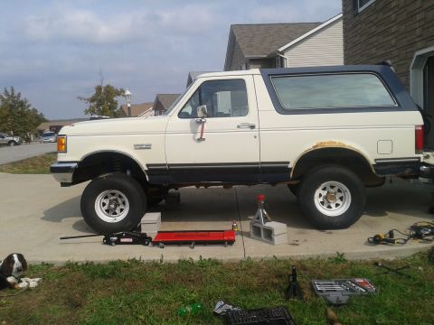 1988 Ford Bronco - A white 1988 Ford Bronco with a blue top is parked on a driveway, lifted for maintenance with various tools around it.