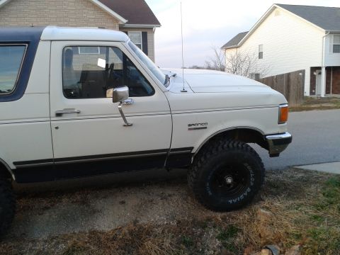 1988 Ford Bronco - Side view of a white Ford Bronco with off-road tires, parked in front of a residential building.