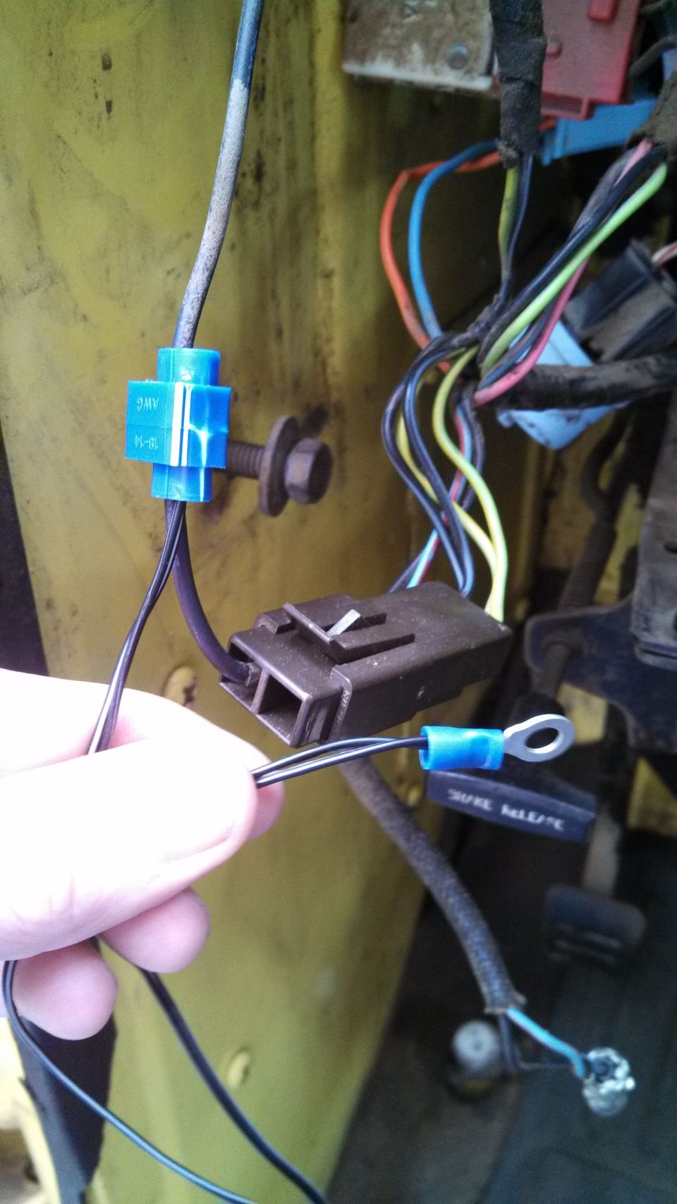 Hand holding black wires with blue quick-splice tap and ring terminal near brown connector on yellow panel