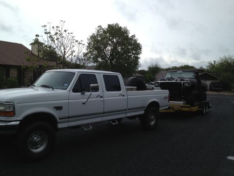 1997 Ford F-350 - A white Ford pickup truck tows a black pickup truck on a trailer under a cloudy sky.
