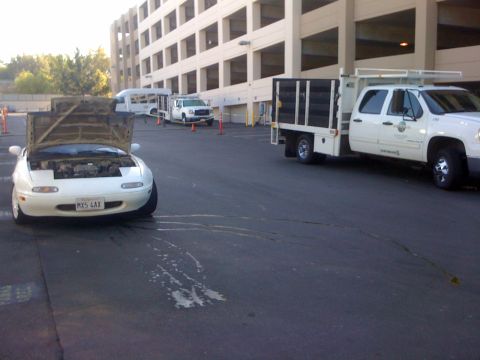 1991 Mazda Miata - A white 1991 Mazda Miata sits with its hood open, revealing the engine, as other vehicles and a parking structure are visible in the backgr…