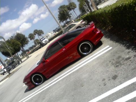1994 Ford Probe - A red coupe is parked in a lot with other vehicles and palm trees under a cloudy sky.