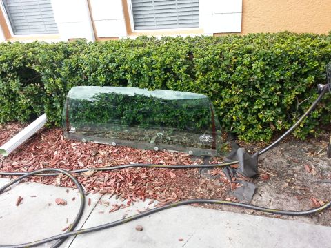 1994 Ford Bronco - curved automotive windshield removed and propped against a hedge near a building, with a garden hose nearby.