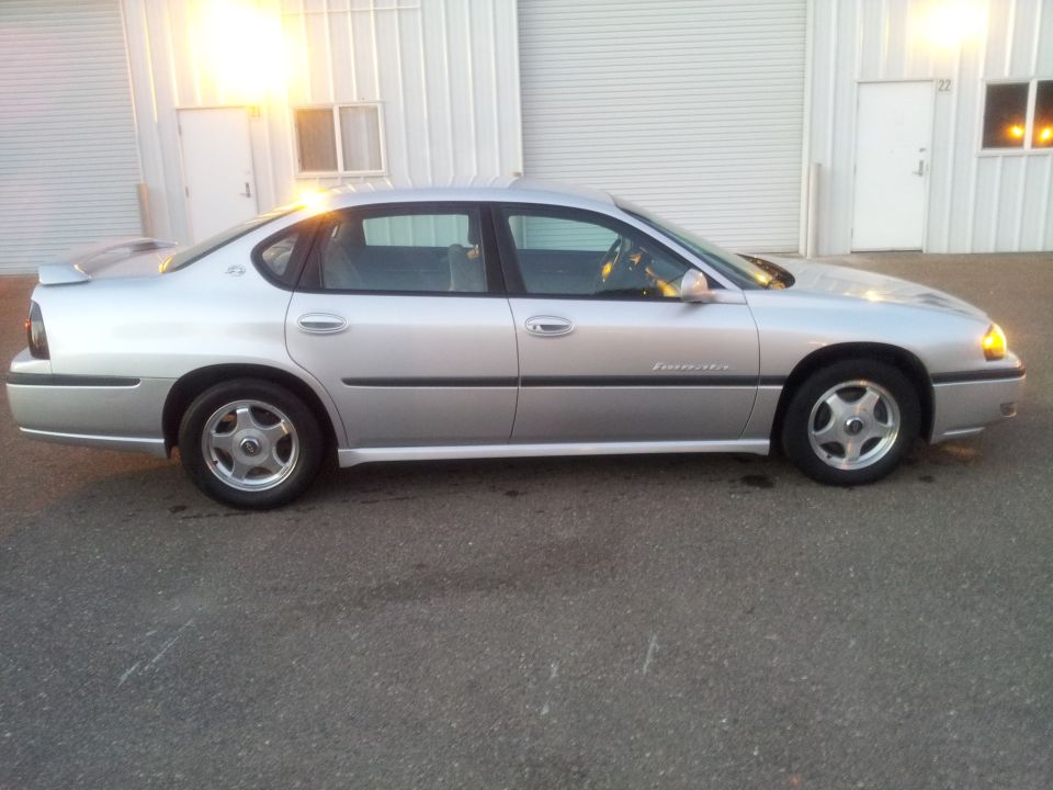 2001 Chevrolet Impala sedan, silver, side view. Parked on asphalt in front of a building with large garage doors