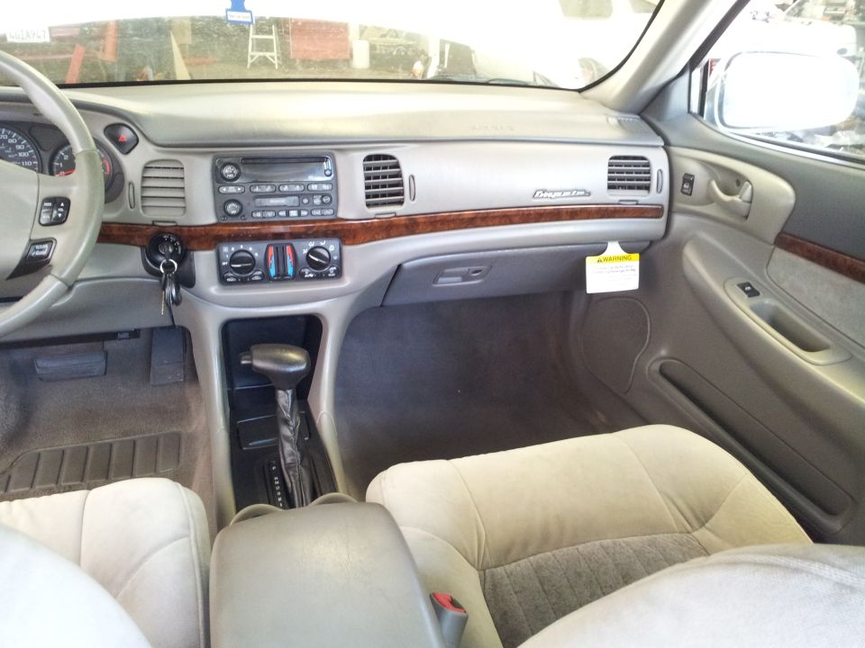 Car interior with beige seats, wood trim dashboard, and automatic gear shifter. A "warning" tag hangs from the glove compartment