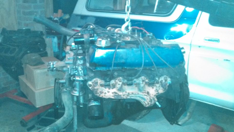 Engine on a hoist with another engine block in the background. A white vehicle is partially visible