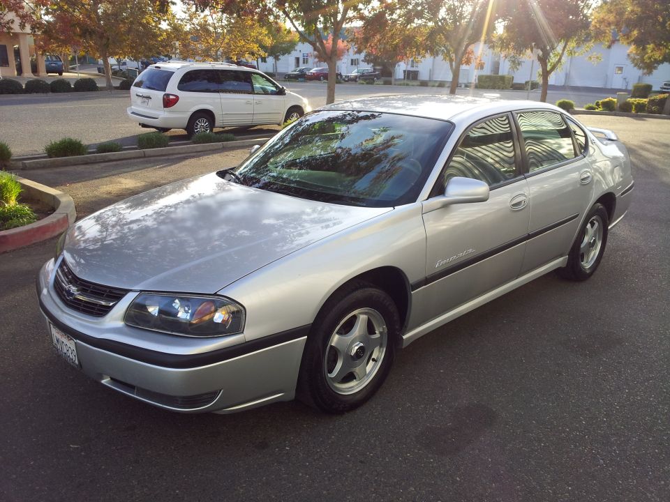 Silver Chevrolet sedan parked on asphalt, white minivan in background
